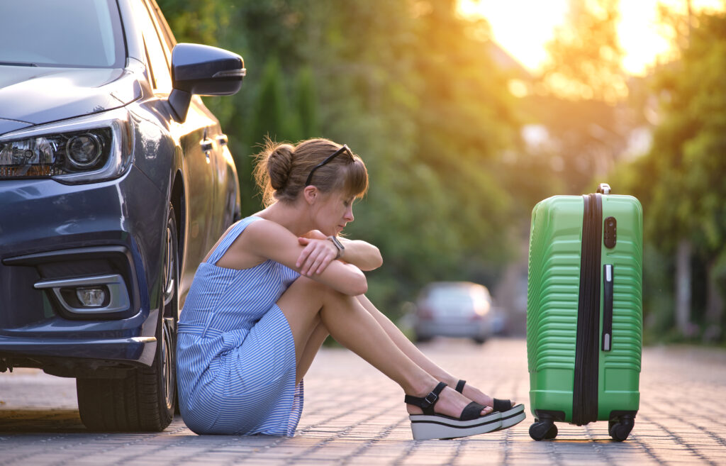 Young tired woman with suitcase sitting beside car waiting for someone. Travelling and vacations concept.
