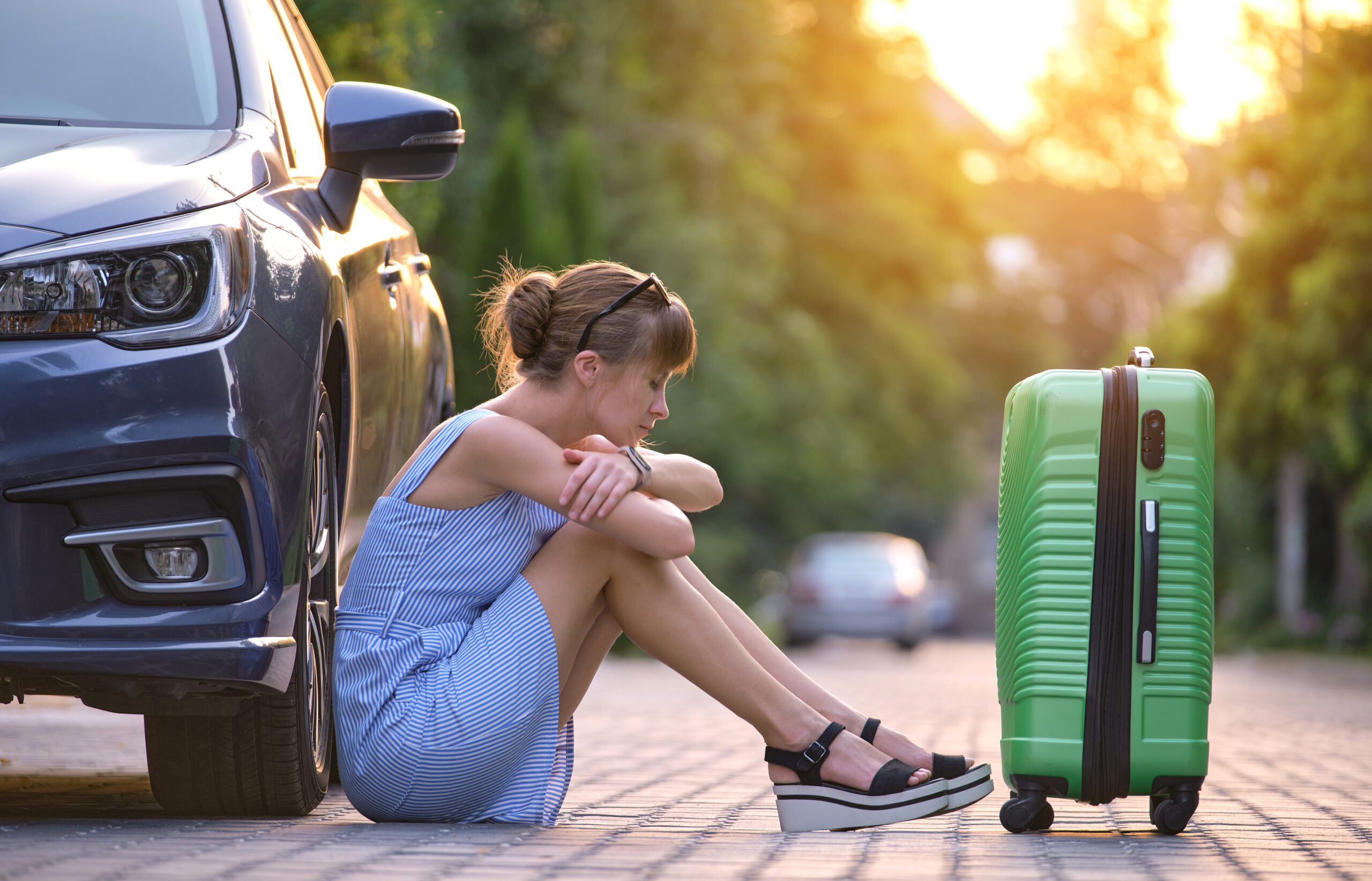 Young tired woman with suitcase sitting beside car waiting for someone. Travelling and vacations concept.